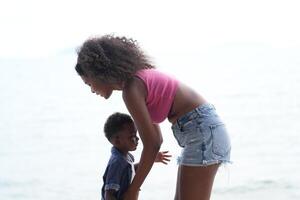 mezclado carrera africano y asiático madre y chico es jugando a el al aire libre área. sonriente contento familia tener divertido corriendo en el playa. retrato de mamá y niño estilo de vida con un único peinado. foto