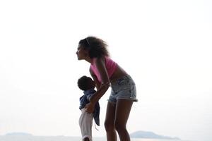 mezclado carrera africano y asiático madre y chico es jugando a el al aire libre área. sonriente contento familia tener divertido corriendo en el playa. retrato de mamá y niño estilo de vida con un único peinado. foto