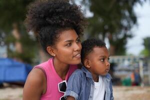 mezclado carrera africano y asiático madre y chico es jugando a el al aire libre área. sonriente contento familia tener divertido corriendo en el playa. retrato de mamá y niño estilo de vida con un único peinado. foto