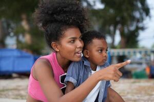 mezclado carrera africano y asiático madre y chico es jugando a el al aire libre área. sonriente contento familia tener divertido corriendo en el playa. retrato de mamá y niño estilo de vida con un único peinado. foto