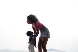 mezclado carrera africano y asiático madre y chico es jugando a el al aire libre área. sonriente contento familia tener divertido corriendo en el playa. retrato de mamá y niño estilo de vida con un único peinado. foto