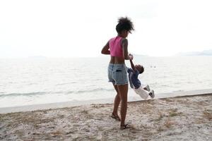 mezclado carrera africano y asiático madre y chico es jugando a el al aire libre área. sonriente contento familia tener divertido corriendo en el playa. retrato de mamá y niño estilo de vida con un único peinado. foto