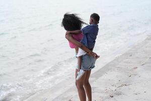 mezclado carrera africano y asiático madre y chico es jugando a el al aire libre área. sonriente contento familia tener divertido corriendo en el playa. retrato de mamá y niño estilo de vida con un único peinado. foto