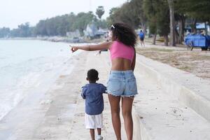 mezclado carrera africano y asiático madre y chico es jugando a el al aire libre área. sonriente contento familia tener divertido corriendo en el playa. retrato de mamá y niño estilo de vida con un único peinado. foto