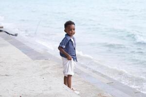 mezclado carrera africano y asiático chico es jugando a el al aire libre área. sonriente contento chico tiene divertido corriendo en el playa. retrato de chico estilo de vida con un único peinado. foto