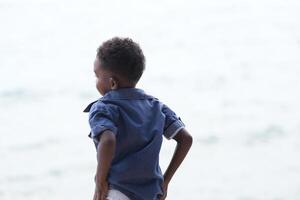 mezclado carrera africano y asiático chico es jugando a el al aire libre área. sonriente contento chico tiene divertido corriendo en el playa. retrato de chico estilo de vida con un único peinado. foto