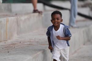 mezclado carrera africano y asiático chico es jugando a el al aire libre área. sonriente contento chico tiene divertido corriendo en el playa. retrato de chico estilo de vida con un único peinado. foto