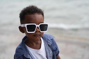 mezclado carrera africano y asiático chico es jugando a el al aire libre área. sonriente contento chico tiene divertido corriendo en el playa. retrato de chico estilo de vida con un único peinado. foto