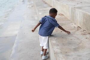 mezclado carrera africano y asiático chico es jugando a el al aire libre área. sonriente contento chico tiene divertido corriendo en el playa. retrato de chico estilo de vida con un único peinado. foto