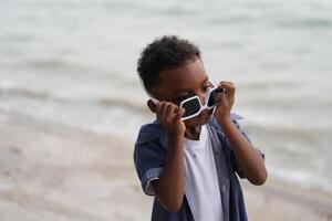 mezclado carrera africano y asiático chico es jugando a el al aire libre área. sonriente contento chico tiene divertido corriendo en el playa. retrato de chico estilo de vida con un único peinado. foto