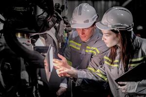 negocio ingenieros reunirse y cheque controlar pesado máquina robot brazo. diverso equipo de industrial robótica ingenieros reunido alrededor máquina. profesional maquinaria operador utilizar industrial digital tableta. foto