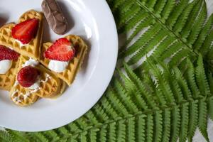 Waffles with strawberry on a plate, green leaves of fern on a table. Close up. Beautiful and tasty breakfast. photo