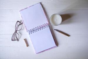 The opened notepad, pen, white candle, glasses and branches of hops as decoration on a white wooden table. Desktop still life with space for text. photo