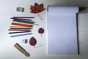 Empty sketch pad, colorful pencils, other equipment for drawing and fallen leaf on the white wooden desktop. Art background with place for text. photo