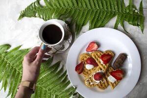 Waffles with strawberry, a cup of coffee and green leaves of fern on a table. Beautiful and tasty breakfast. photo