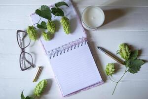 The opened notepad, pen, white candle, glasses and branches of hops as decoration on a white wooden table. Desktop still life with space for text. photo