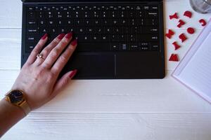 Female hand with red manicure on a keyboard. Laptop, notepad and red letters on a white wooden desktop. photo