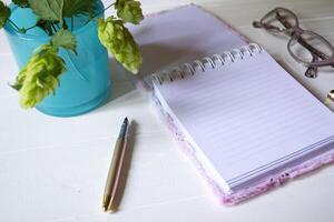 The opened notepad, pen, white candle, glasses and branches of hops as decoration on a white wooden table. Desktop still life with space for text. photo