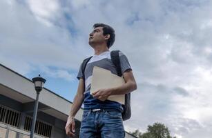 un masculino estudiante participación un archivo y mirando arriba a el cielo foto