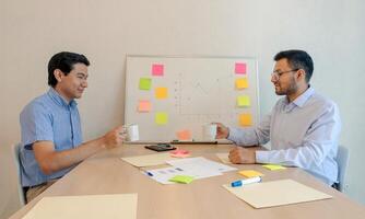 Two men sitting at a table with a whiteboard and a white board with post it notes photo