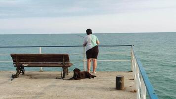 A woman standing fishing in the sea next to a bench with her dog video