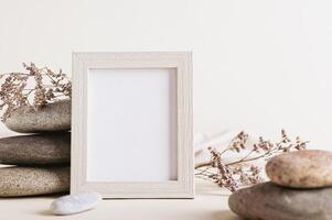 An empty photo frame stands near a stack of stones and dried flowers on a light background