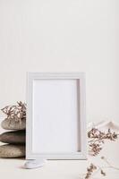 An empty photo frame near a stack of stones and dried flowers on a light background vertical view