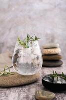 Ginebra tónico con hielo y Romero en un vaso en un Roca en el mesa vertical ver foto