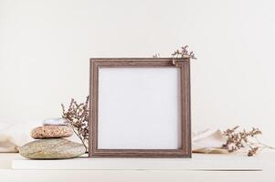 Square empty photo frame surrounded by dried flowers and stones on a light beige background