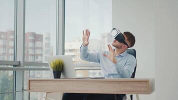 A young man sitting at a desk in the office uses augmented reality glasses to work on business projects in various fields. video