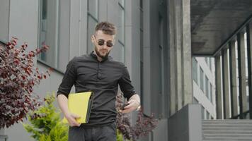 affärsman med glasögon och en gul mapp med dokument väntar för en möte och ser på hans Kolla på. man väntar för en företag möte på de bakgrund av modern byggnader i de stad Centrum. video