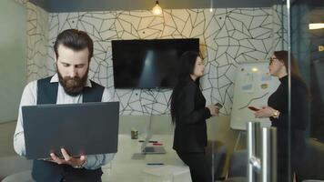 ufficio opera. uomo con un' barba Tenere un' il computer portatile nel il suo mano e Lavorando. Due donne nel il sfondo siamo parlando. video