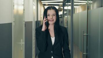 de cerca positivo negocio mujer hablando teléfono inteligente en oficina interior. sonriente mujer de negocios vocación móvil teléfono en corredor. retrato de simpático dama caminando en negocio centrar pasillo. video