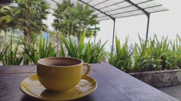 geel keramisch koffie kop Aan een cafe tafel, bokeh achtergrond van groen bomen zwaaiend in de wind video