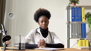 africano americano fêmea médico olhando às Câmera enquanto sentado às mesa dentro hospital video