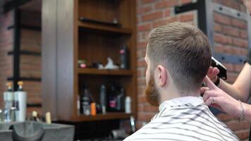 A young barber makes a trimmer haircut of a client with a red beard. Back view video