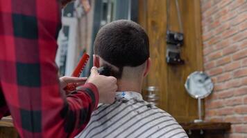 jovem à moda cabeleireiro dentro uma xadrez vermelho e Preto camisa faz uma corte de cabelo em a costas do a do cliente cabeça dentro uma barbearia video