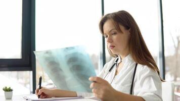 Young caucasian woman doctor wearing white medical coat and stethoscope writes a medical history in a notebook and holds the x-ray video