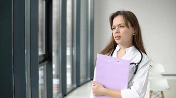 Happy young caucasian woman doctor wearing white medical coat and stethoscope with documents in her hands looks to the side. Smiling female physician posing in hospital office video