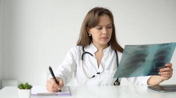 Young caucasian woman doctor wearing white medical coat and stethoscope writes a medical history in a notebook and holds the x-ray video