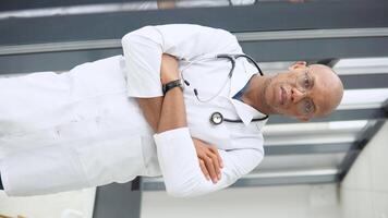 A young african american male doctor stands in a medical suit at the clinic and looks at the camera. Vertical video