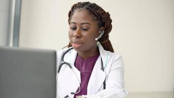 joven africano americano mujer médico con auriculares teniendo charla o consulta en ordenador portátil video