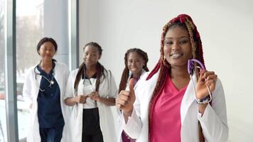 A young african american doctor with red hair stands in front and looks at the camera against the background of a team of colleagues video