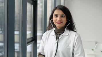 Happy young indian woman doctor in white medical coat wears a stethoscope and looking at camera. Smiling female physician posing in hospital office video