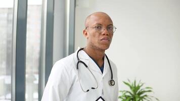 A young african american male doctor stands in a medical suit at the clinic and looks at the camera video