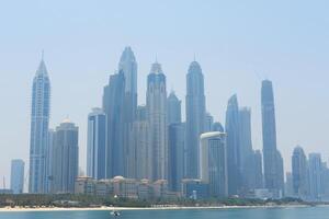 A hazy view of Dubai's skyscraper-filled skyline with beachfront and the Persian Gulf in the foreground. Dubai, UAE - August 15, 2023 photo