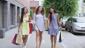 Three beautiful young girlfriends are walking along the street with packages after shopping and discussing purchases. slow motion. HD video