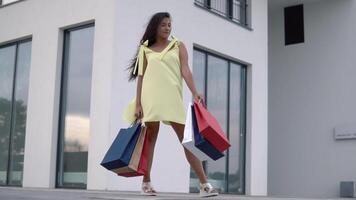 schön Mädchen Modell- im ein lange Gelb Kleid nach Einkaufen mit farbig Taschen im Hände haben ein gut Stimmung auf ein Weiß Gebäude Hintergrund. schleppend Bewegung. video