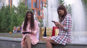 zwei schön jung Mädchen nach Einkaufen sind Sitzung in der Nähe von das Brunnen im das Park. schleppend Bewegung. hd. video