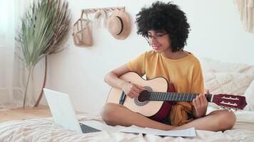 jugando guitarra, en línea yo estudiar, Arte lecciones, utilizar un artilugio. afro americano niña en un amarillo camiseta aprende a jugar el guitarra utilizando un ordenador portátil. video